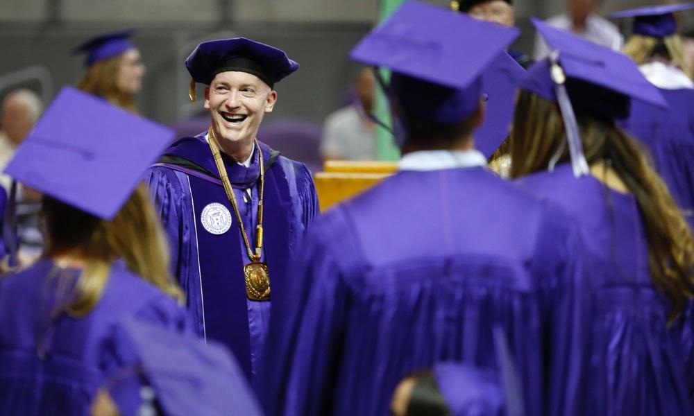 Chancellor Victor J. Boschini, Jr., smiling and in full academic regalia, greeting TCU graduates in purple caps and gowns